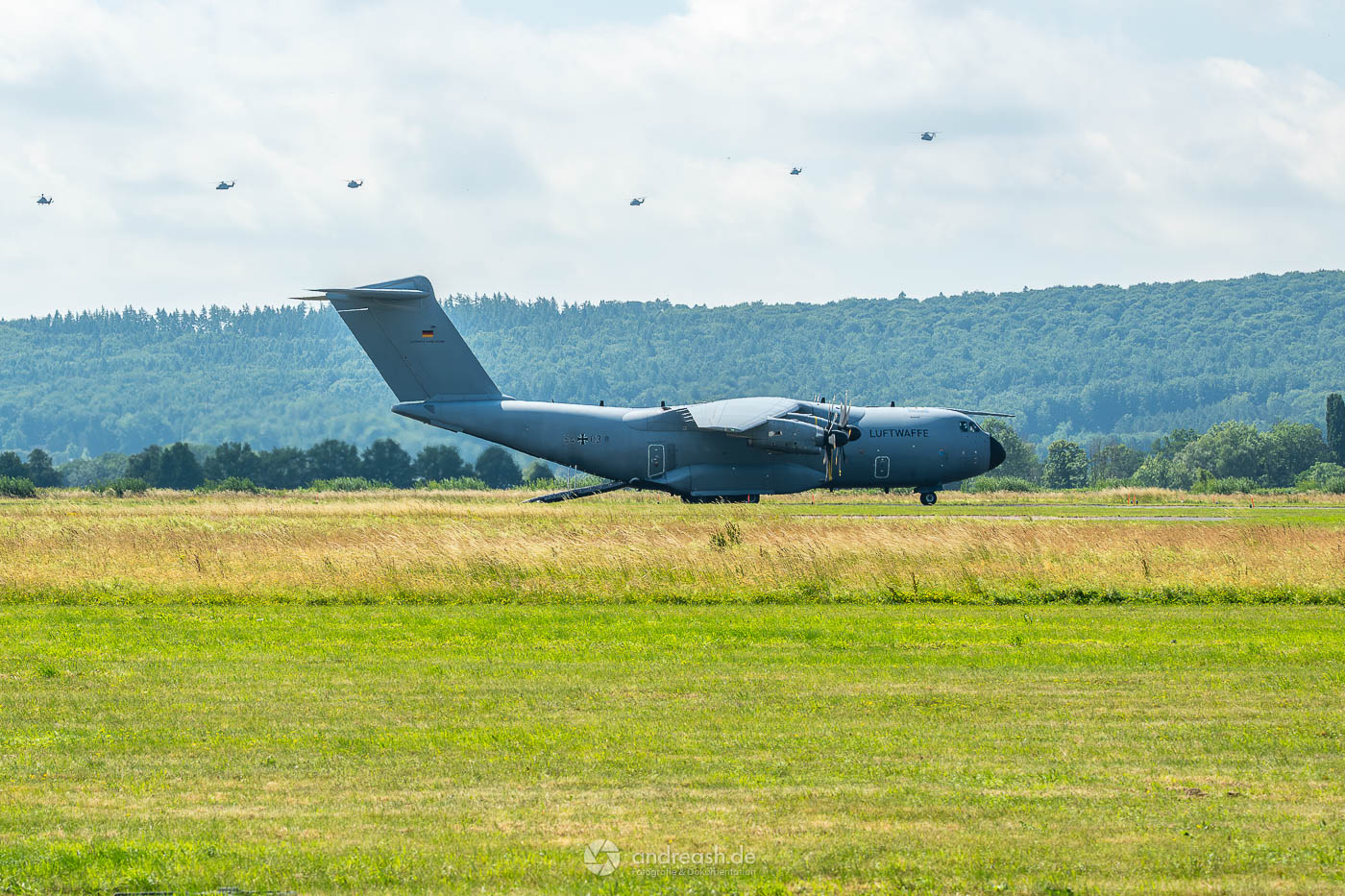 Eine A400M der Luftwaffe steht mit laufenden Turbinen und geöffneter Heckrampe auf dem Rollfeld. Im Hintergrund formieren sich Kampfhubschrauber Tiger, NH90 und eine CH-53G für den Abschlussvorbeiflug.
