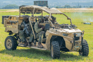 Fallschirmjäger sitzen in einem leichten luftlandefähigen Utility Terrain Vehicle (LL UTV) und überwachen das Gelände, im Hintergrund gelber Rauch.